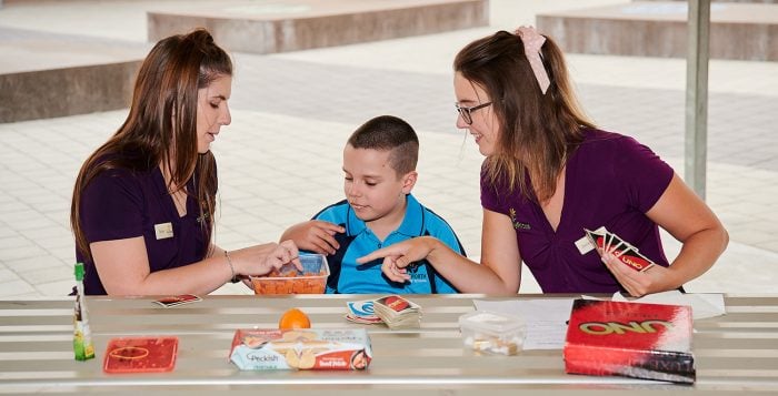 two therapists show food to boy. They are sitting at a metal picnic table. 