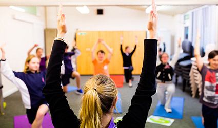 Students and therapist engage in Yoga. They stand on mats with their arms raised in the air.
