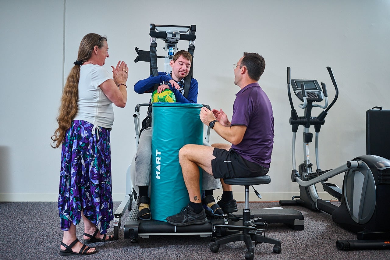 A young man stands in a Lite Gait on a treadmill. He stands behind a circular foam pillar and holds a volley ball. His mum stands next to him cheering. They are joined by their physiotherapist who is clapping and sitting on a stool.