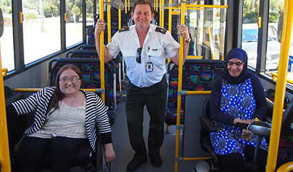 A transport officer stands in a bus between two women who use wheelchairs.