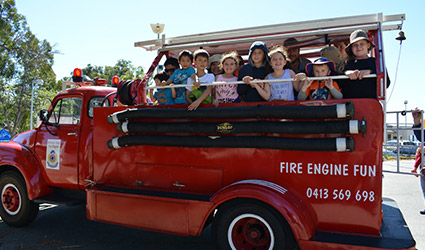 A group of children smile while standing on the back of a fire engine.