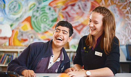 Jayden and his therapist sit at a desk in a classroom as they laugh.