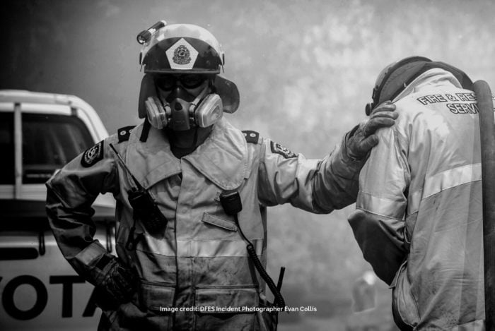 Two firefighters stand together in full uniform and wearing a breathing apparatus. One firefighter is facing away from the camera with their head in their hands. The other firefighter is facing towards the camera and has on hand on his hip and one hand on the other firefighter shoulder. The image is in black and white.