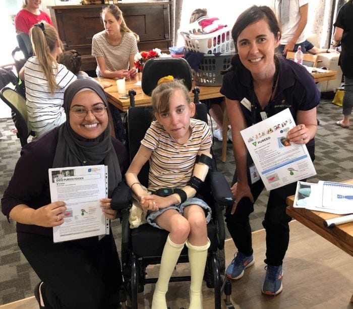 Zahraa (left) crouches next to Leana (middle) and Kim (right) stands next to Leana. They are all looking at the camera smiling. Leana uses her wheelchair and wears a stripped t-shirt. Zahraa and Kim wear their Therapy Focus uniform.
