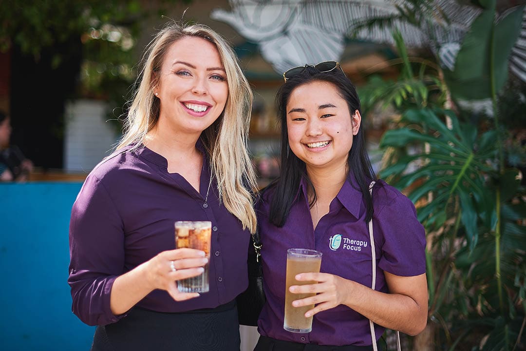 Two graduate therapists stand together and smile at the camera. They hold drinks and are wearing purple Therapy Focus tops.