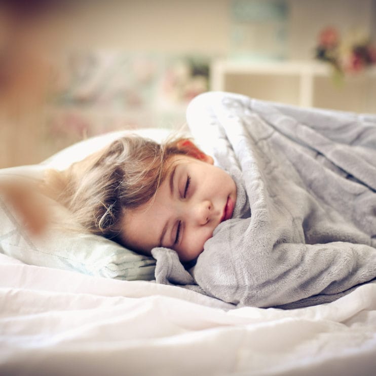 A child lays in bed with a weighted blanket over them. They are peacefully sleeping.