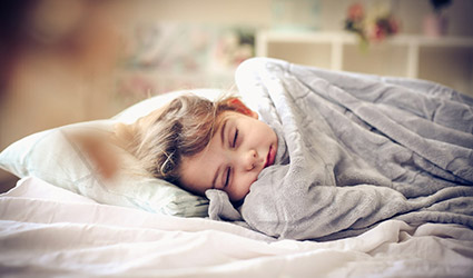 A child lays in bed with a weighted blanket over them. They are peacefully sleeping.