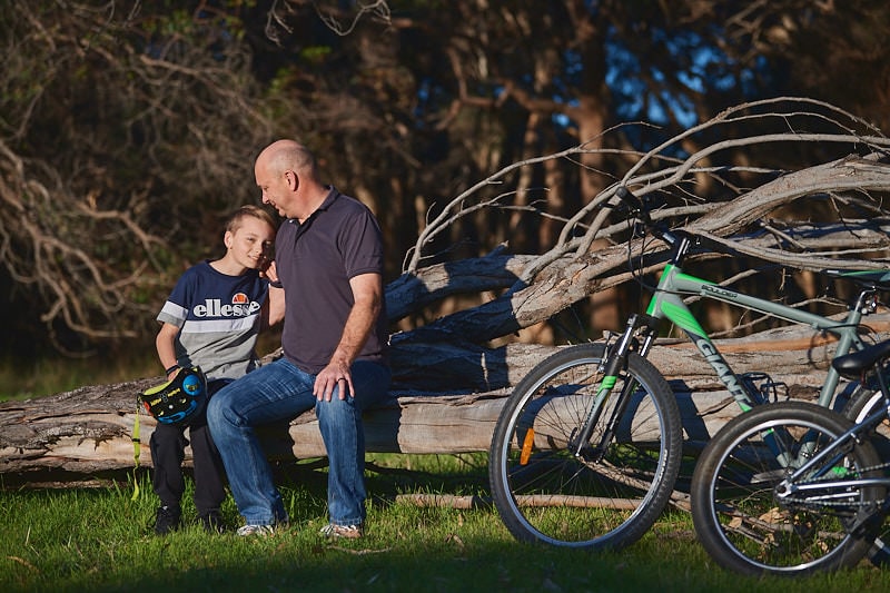A father and son sit on a fallen tree and embrace. A green bike sits to the right of them.