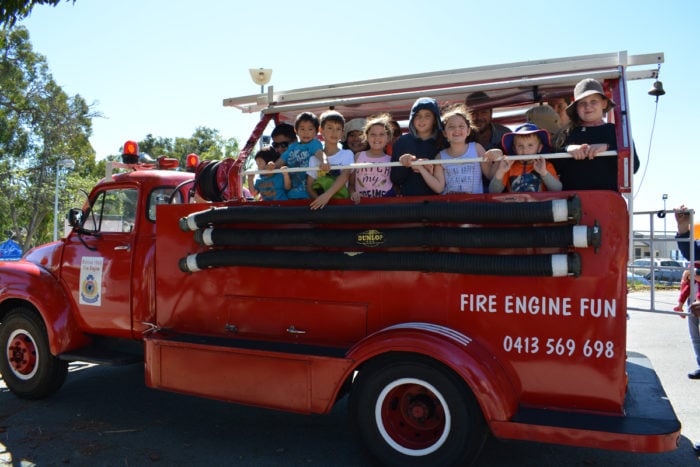 A group of children smile while standing on the back of a fire engine. 