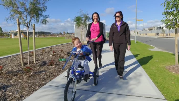 Theodore rides his bike and is accompanied by mum Sharon and his therapist. They are moving through a park on a pathway. 