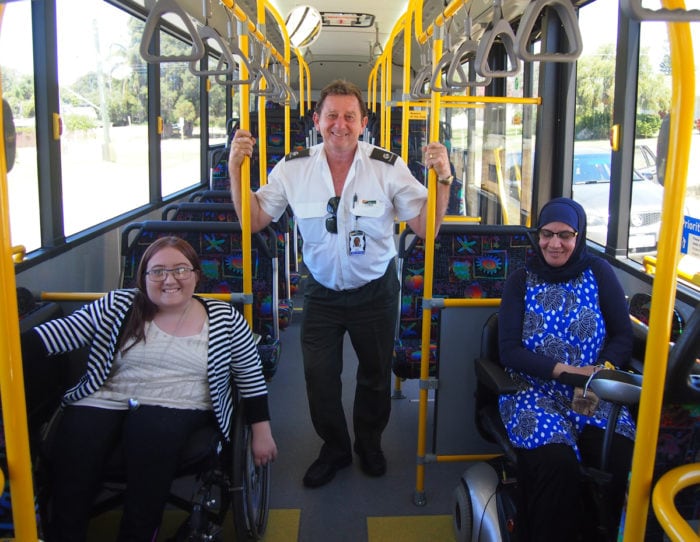 A transport officer stands in a bus between two women who use wheelchairs.
