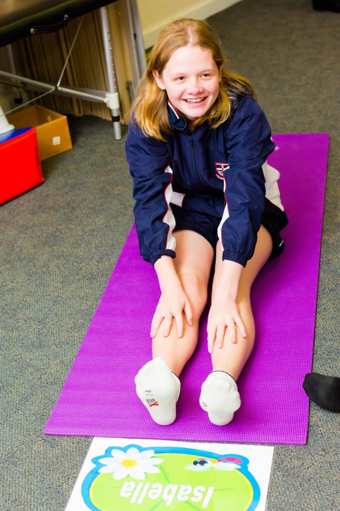 A young girl sits on a pink yoga mat and smiles. She stretches her hands towards her legs in front of her. 