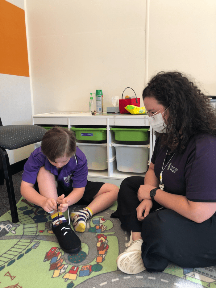 Cameron sits on the mat next to Maisie and ties his shoelaces. He is looking intently at his sneaker in front of him.