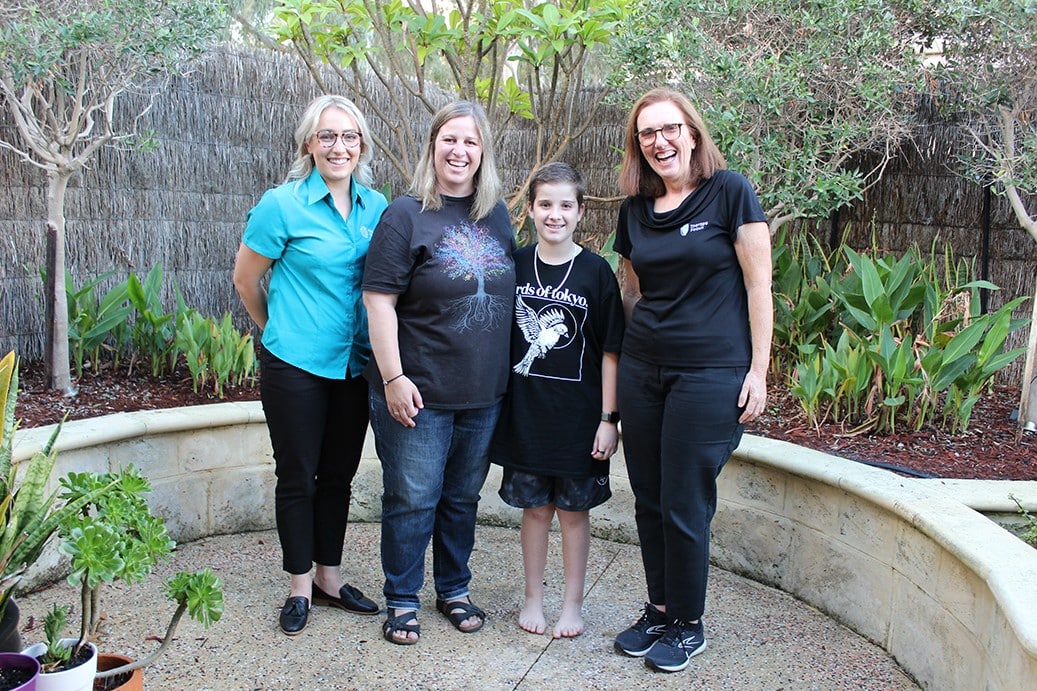 Two therapists, a young boy and a mother stand in a garden next to each other. They are smiling towards a camera.