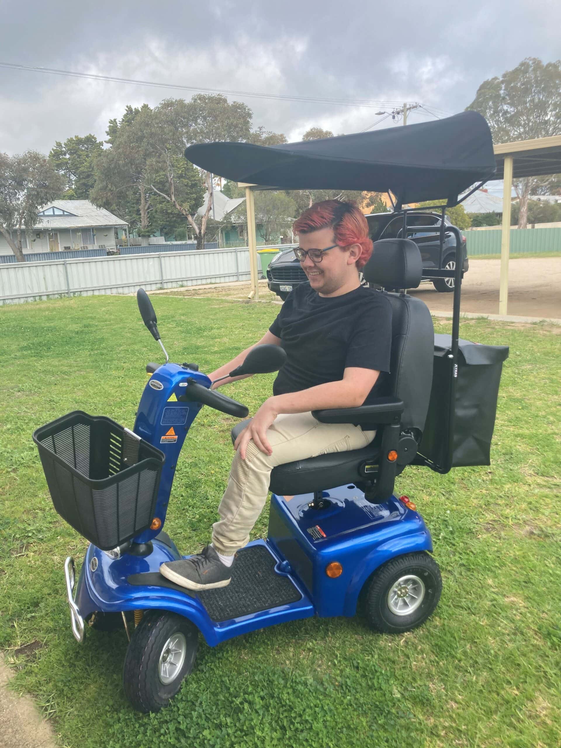 A young man sits on a blue scooter. He has red hair and wears a black t-shirt and cream pants. His scooter has a black basket on the front and a black shade over the top. He is parked on a lawn.
