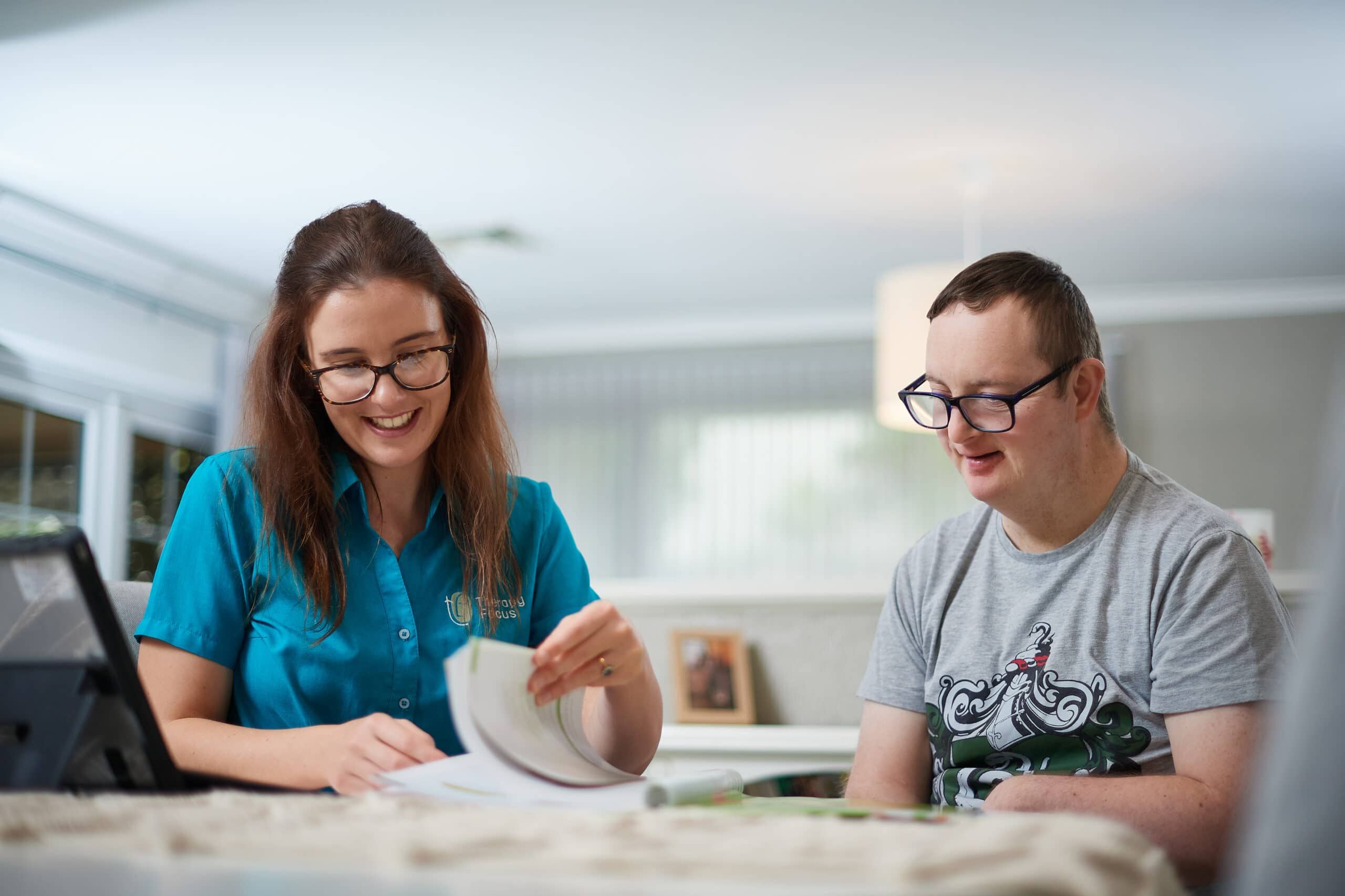 A therapist and young adult sit at a table and examine a booklet. Ther therapist is wearing a blue top and has medium length brown hair. The young adult wears glasses and a grey top. 
