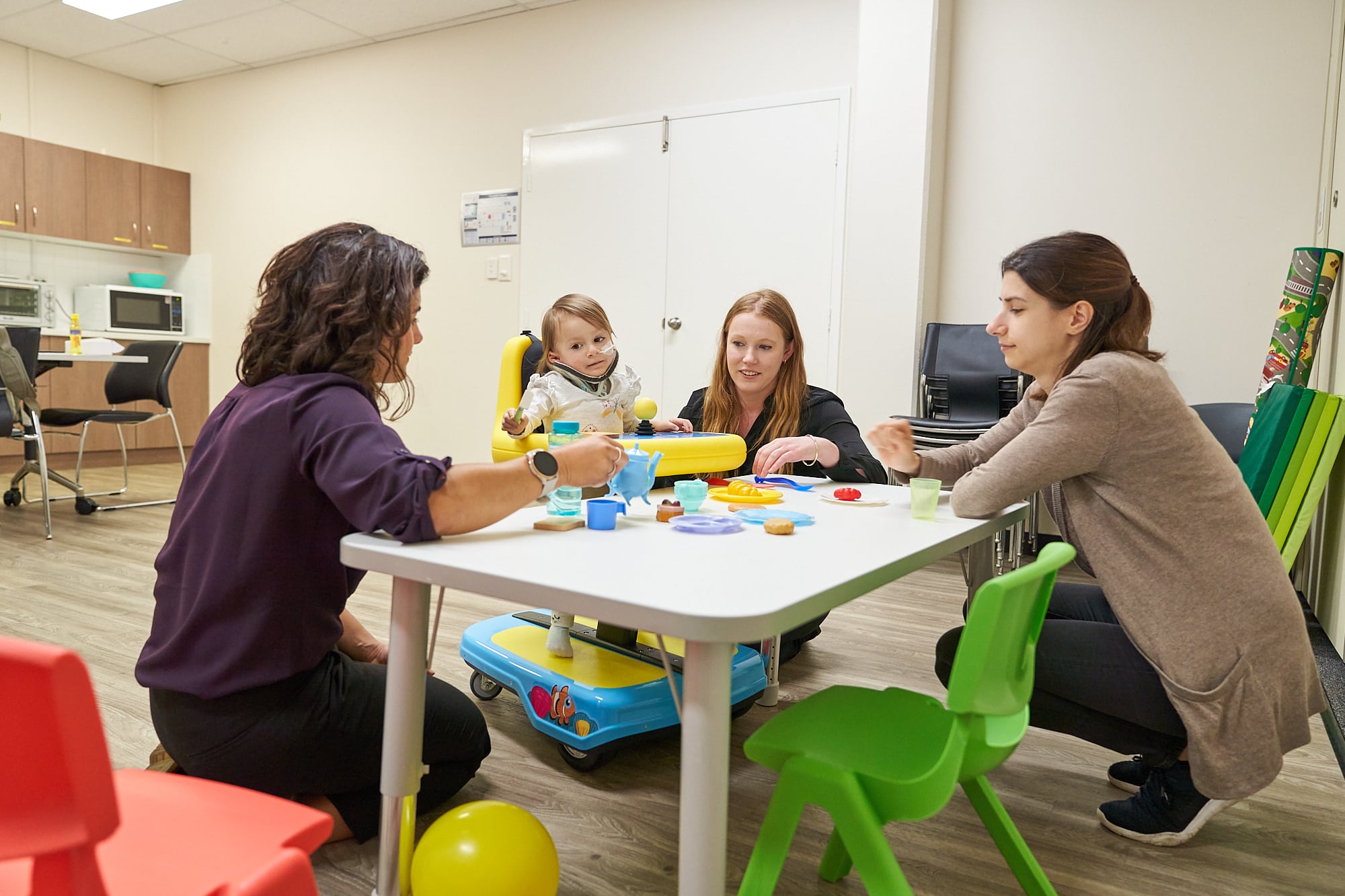 A therapist supports a young child as she engages in play. They are sitting at an activity table with two other women. They are playing with a teapot and cup set. 