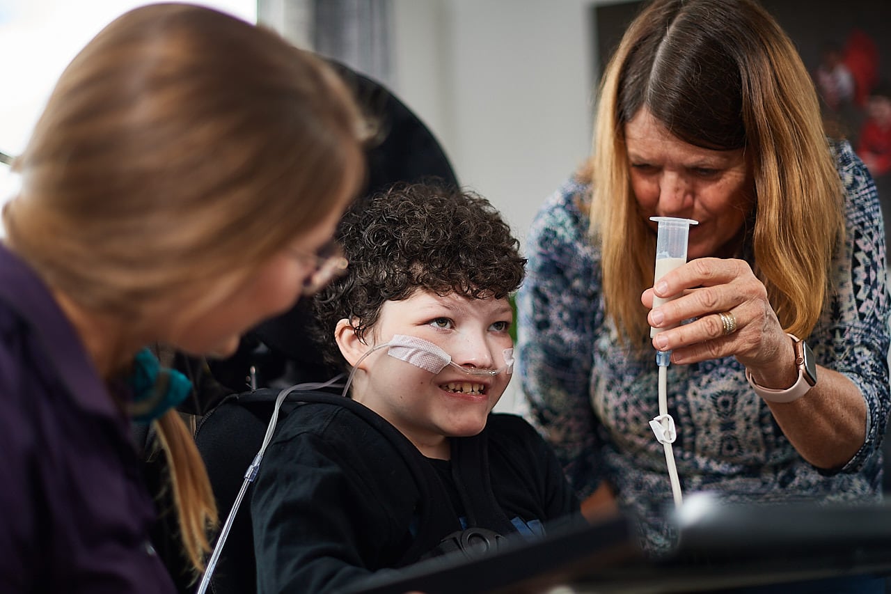 Young boy smiles, his mother is holding a feeding tube and his therapist is next to him.