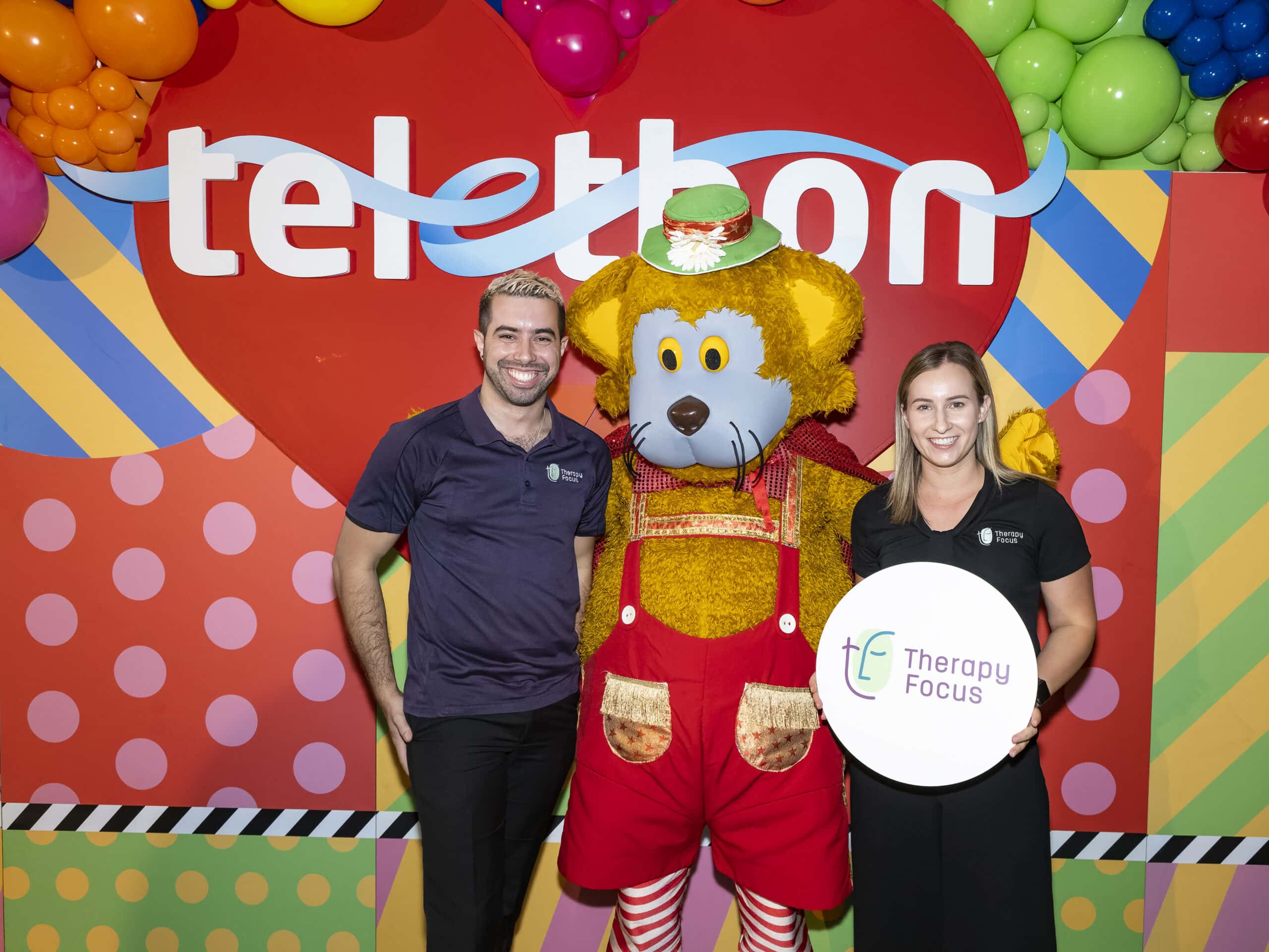A man and woman stand either side of Fat Cat against a colourful wall that says Telethon. The man had brown and blonde hair and wears a purple Therapy Focus polo shirt. The woman has mid-length light hair and wears a black Therapy Focus shirt.