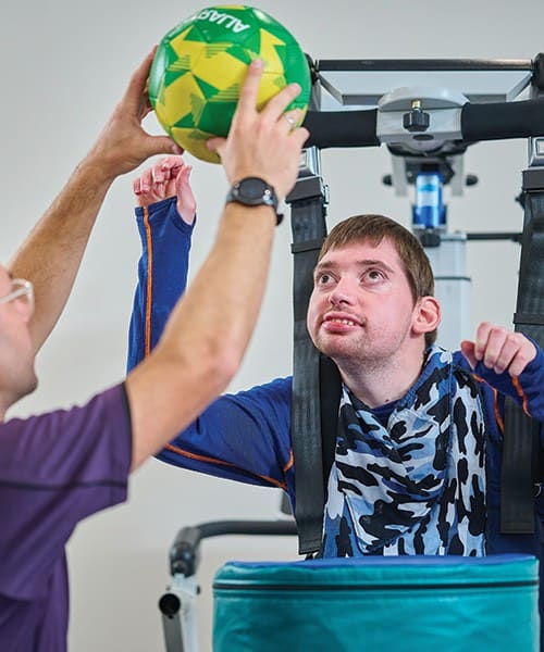 A young man in a Litegait harness reaching for a ball his therapist is holding