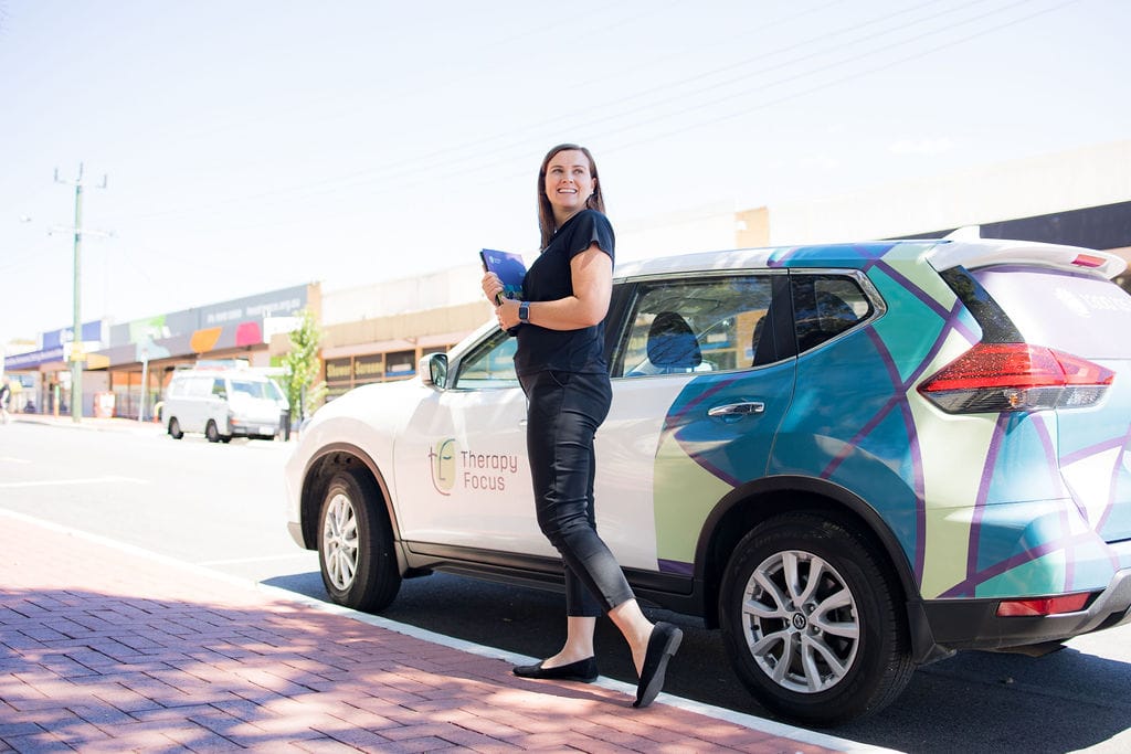 A therapist walks towards her car. She holds a folder and is looking back over her shoulder. She has brown mid-length hair and wears a black uniform.